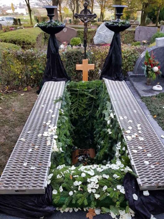 Ausgehobenes Grab mit Innenverkleidung aus Tannenzweigen dekoriert mit weißen Blütenblättern